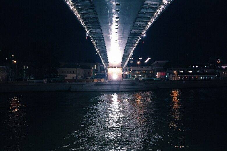 The illuminated underside of Patriarshy Bridge above the dark waters of the Moskva River; the embankment and buildings are visible on the far bank.