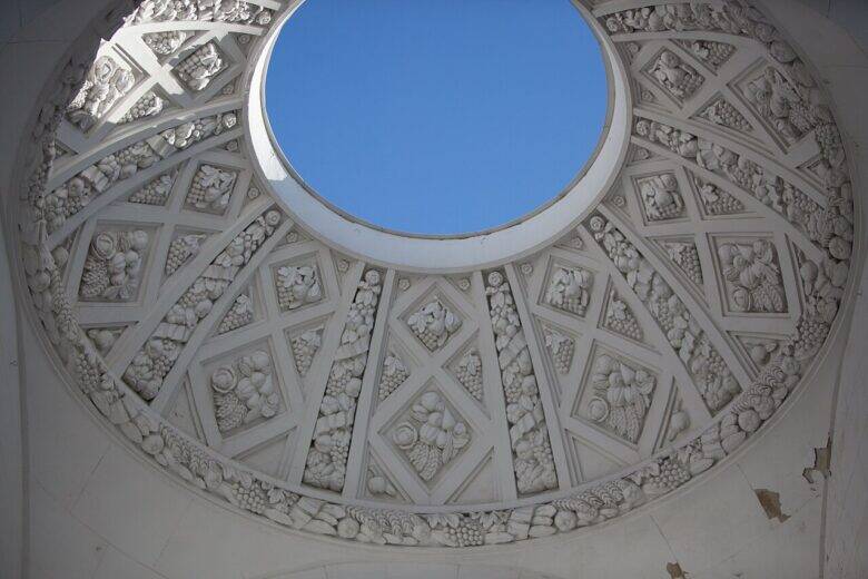 A circular opening revealing blue sky in the centre of a white ceiling, surrounded by radial sections decorated with botanical reliefs.