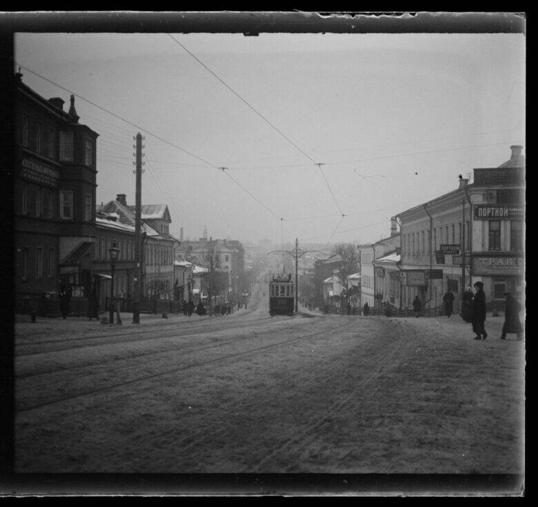 A black-and-white photograph of a street with a tram in the centre, tracks in the foreground, wires above the road and pedestrians beside the buildings.