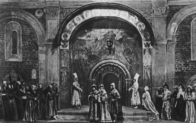 A black-and-white photograph of a scene from Tsar Fyodor Ioannovich: actors in historical costumes in front of a large painted arch.