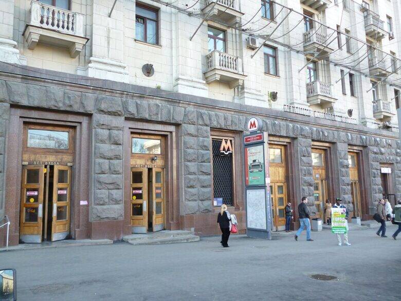 Metro entrance doors marked with an ‘M’ in the high-rise’s stone-clad ground floor; balconies are visible above, with pedestrians walking past the façade.
