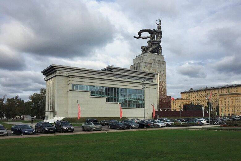 The ‘Worker and Collective Farm Woman’ sculpture atop a tall pavilion, with cars and a lawn in front.