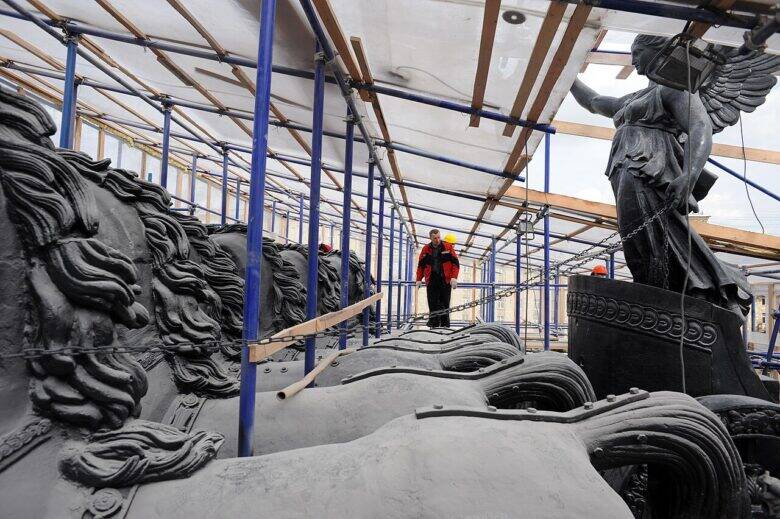 Скульптуры коней и крылатой фигуры среди строительных лесов; между ними стоит рабочий.
