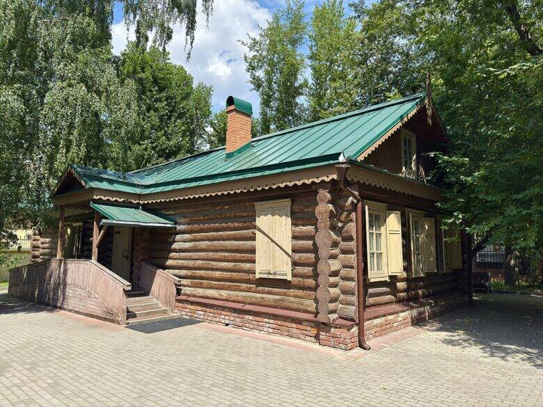 Угол бревенчатой Кутузовской избы с зелёной крышей, светлыми ставнями и входом.