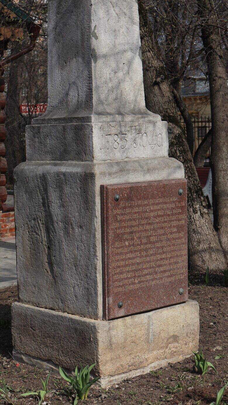 Каменный столб с надписью «1783 года» над красноватой памятной доской; позади деревья.
