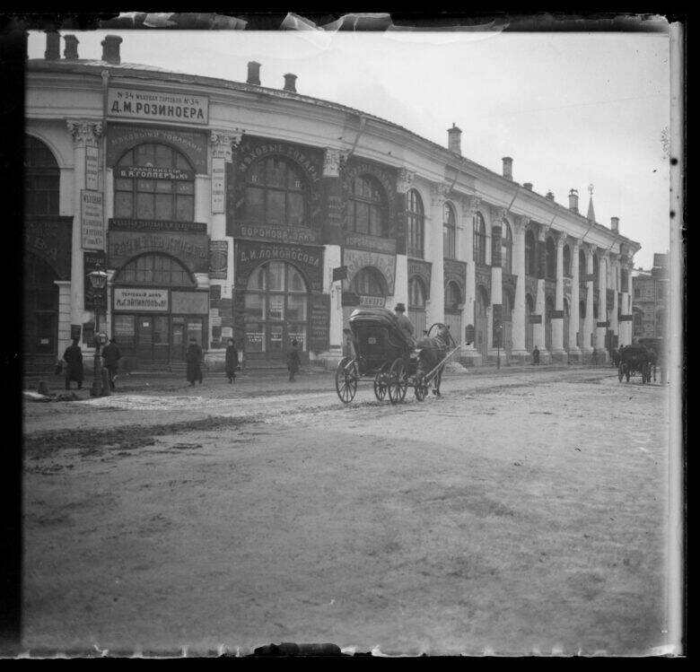Чёрно-белая фотография длинного арочного фасада Старого Гостиного двора; на улице стоят люди и конный экипаж.