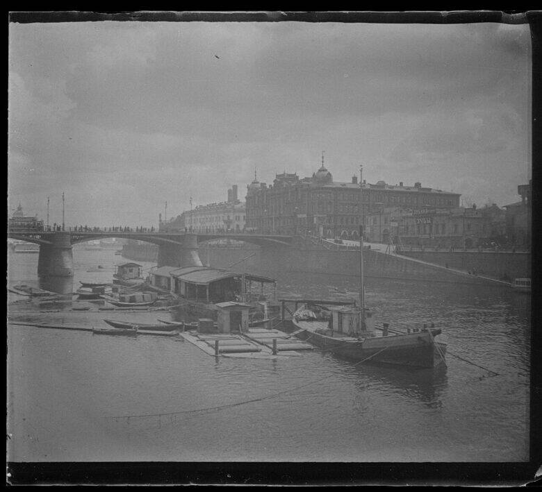 An old bridge across the Moskva River, with boats and floating structures in front of it and city buildings beyond.