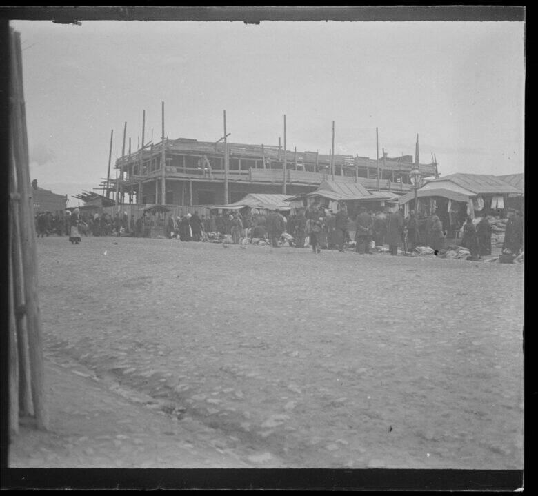 A black-and-white photograph: people beside market awnings and goods laid out for sale; a building covered in scaffolding behind them and a cobbled square in the foreground.