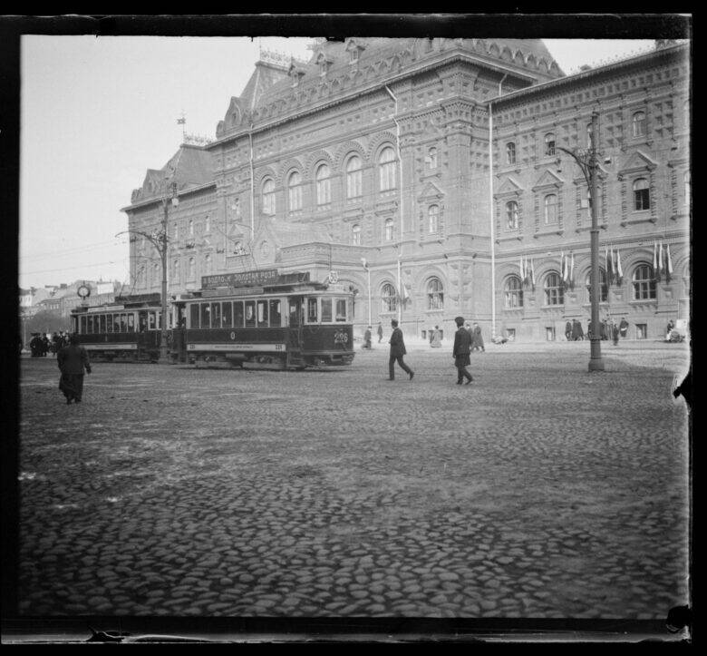 A tram and pedestrians on a paved square in front of a building with tall arched windows.