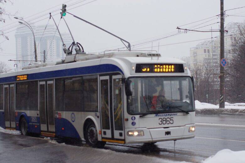 一辆车身编号为3865的蓝白色无轨电车，在架空接触线下沿积雪的街道行驶。