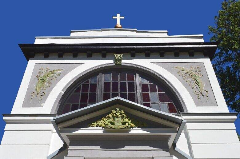 The upper part of the mausoleum’s pale façade, with an arched window, decorative reliefs and a cross on the roof.