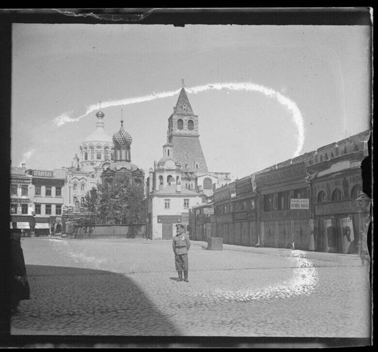An archival photograph of a cobbled square: a person stands in front of a gate tower, with church domes visible on the left and buildings with signs on the right.