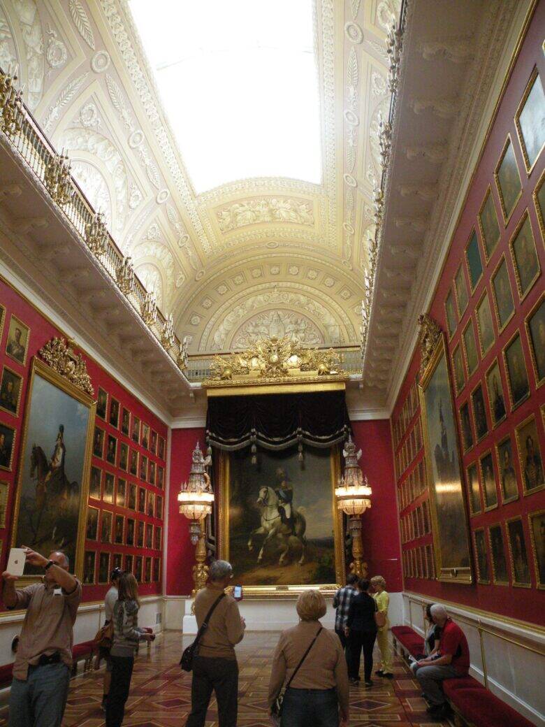 A hall with red walls, rows of small portraits, a high vaulted ceiling and a large painting at the far end; visitors in the foreground.