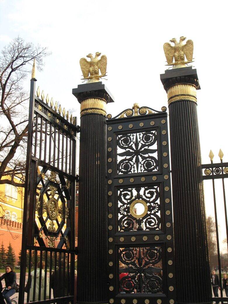 Gates with black fluted pillars, gilded double-headed eagles and decorative openwork ironwork; a red wall behind them.